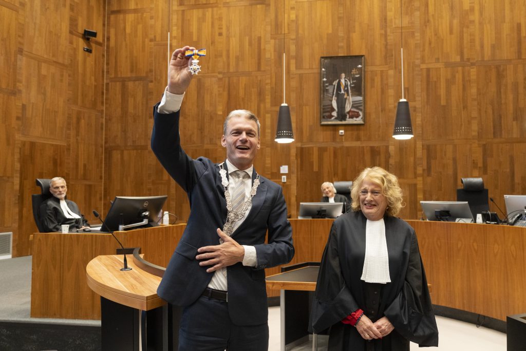 man steekt hand omhoog en houd onderscheiding vast. Vrouw staat er lachend naast.