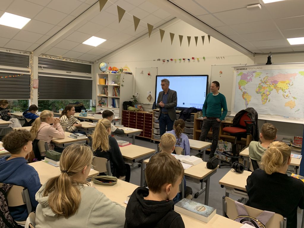 Wethouder Albert Kerssies en een ambtenaar van de gemeente staan voor de klas. De leerlingen luisteren naar hen. 