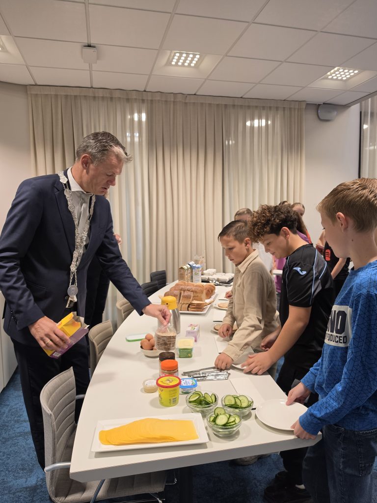 Kinderen staan aan een ontbijtbuffet en kijken naar eten. De burgemeester wijst aan wat er ligt.